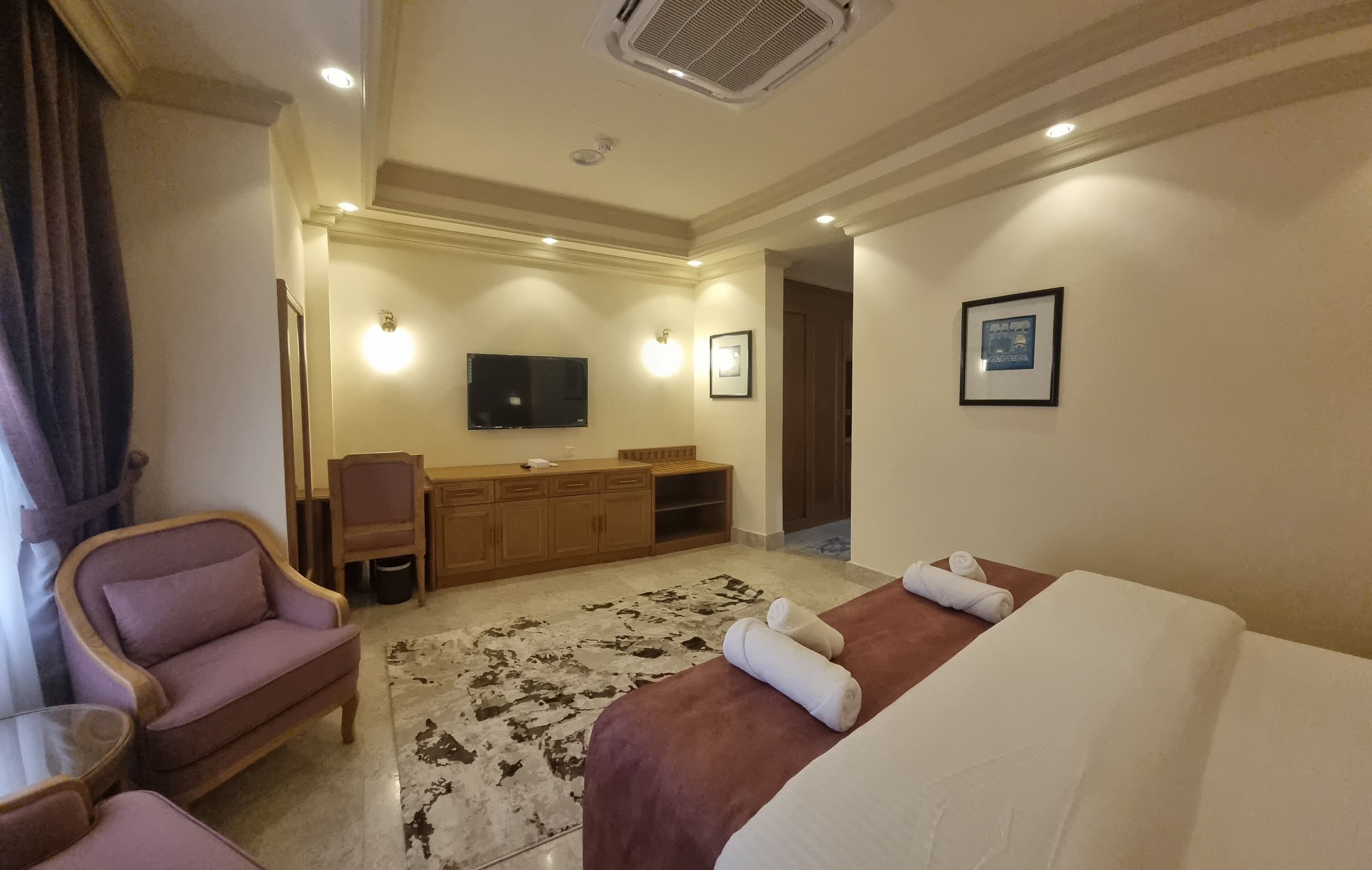 Habitación de hotel con cama, zona de estar con una silla, escritorio, televisión y baño visible a través de una puerta abierta.