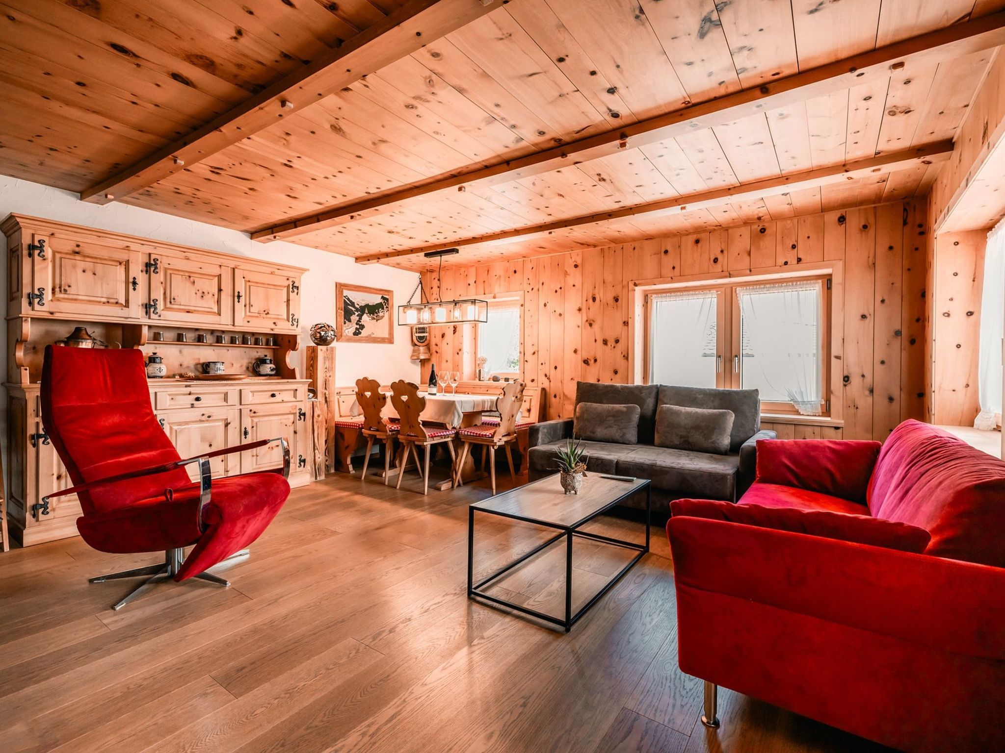 Ein gemütliches Blockhaushaus-Interieur mit Holzwänden und -decken, einem Essbereich mit Tisch und Stühlen sowie einem Wohnzimmer mit einer Couch und einem roten Sessel.