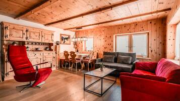 A cozy cabin interior with wooden walls and ceilings, a dining area with a table and chairs, and a living room with a sofa and a red chair.