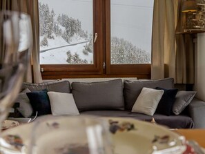 A living room with a sofa, a window with a view of a snowy landscape, and a wooden door. - Spacious apartment, panoramic view - 6 guests (La Plagne)