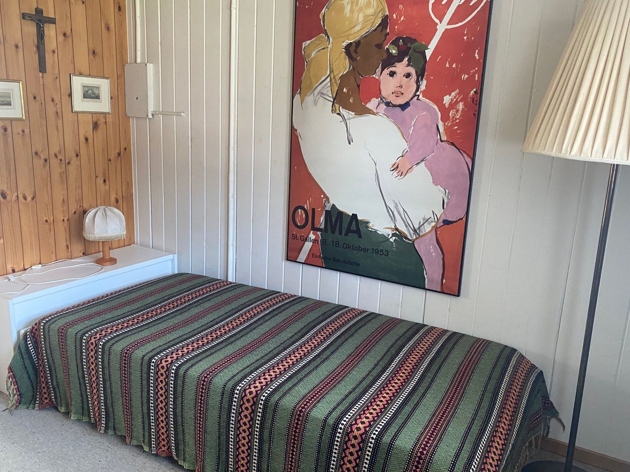 Ein Zimmer mit einem Bett, einer Lampe, einem Poster und einer Holzwand mit einem Kreuz.
