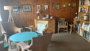 Ein Zimmer mit einem runden Tisch, Korbstühlen, einem Bücherregal und gerahmten Bildern an der Wand.