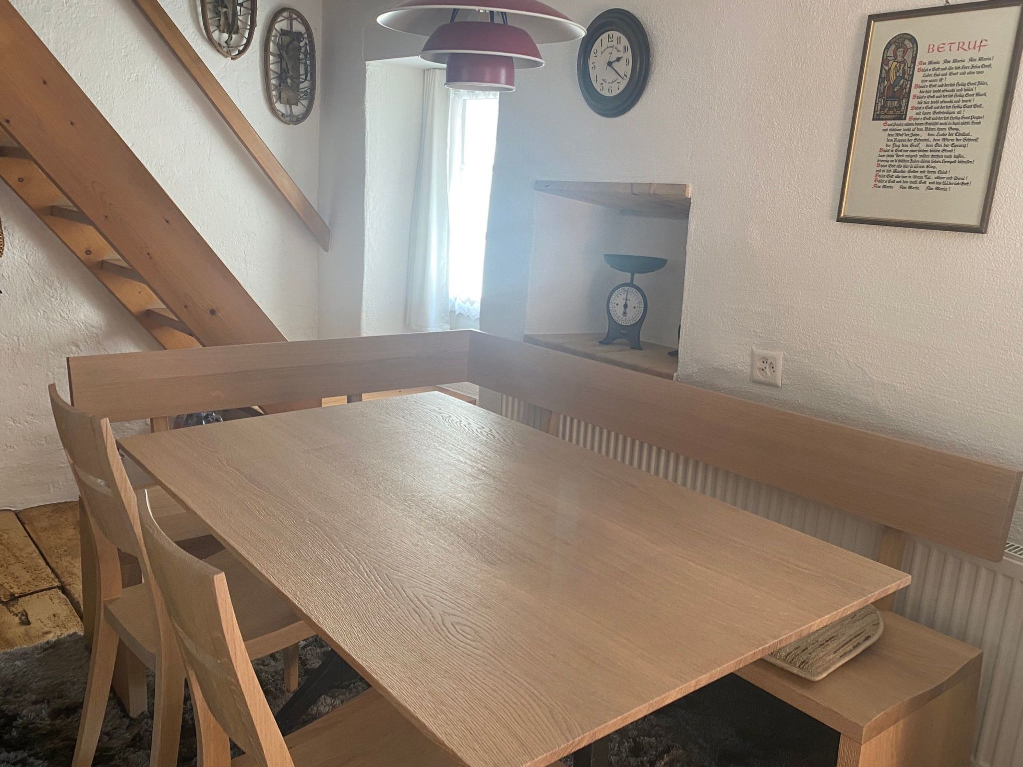 Ein Eckzimmer mit Holztisch und Bänken, einer Treppe und einer Wanduhr.