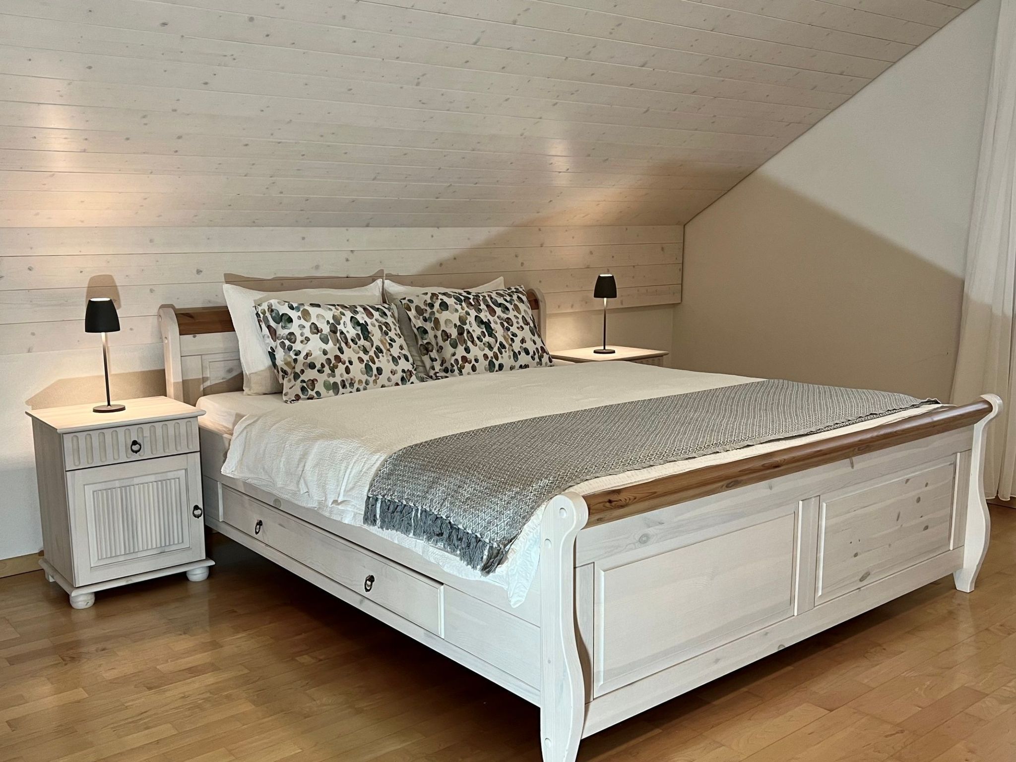 Une chambre avec un plafond en pente, un lit doté d’une tête de lit en bois, deux tables de chevet avec des lampes et un plancher en bois.