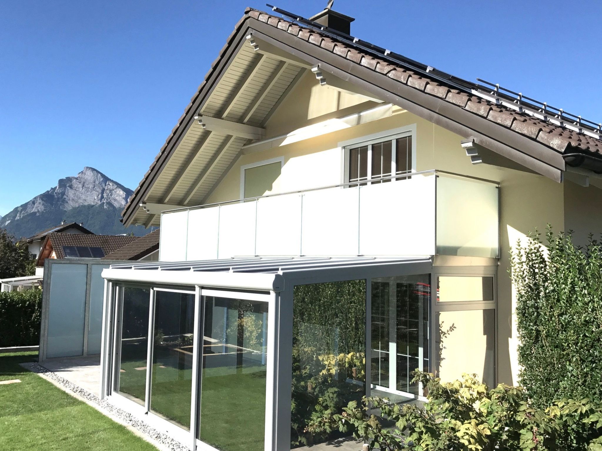 Une maison moderne à deux étages, dotée d’une véranda en verre, avec une vue sur les montagnes et un jardin bien entretenu.