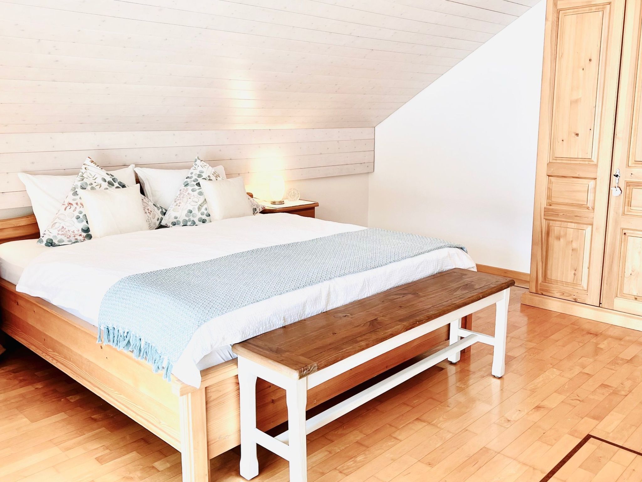 Een slaapkamer met een houten bed, wit beddengoed, een bankje en een houten garderobe.