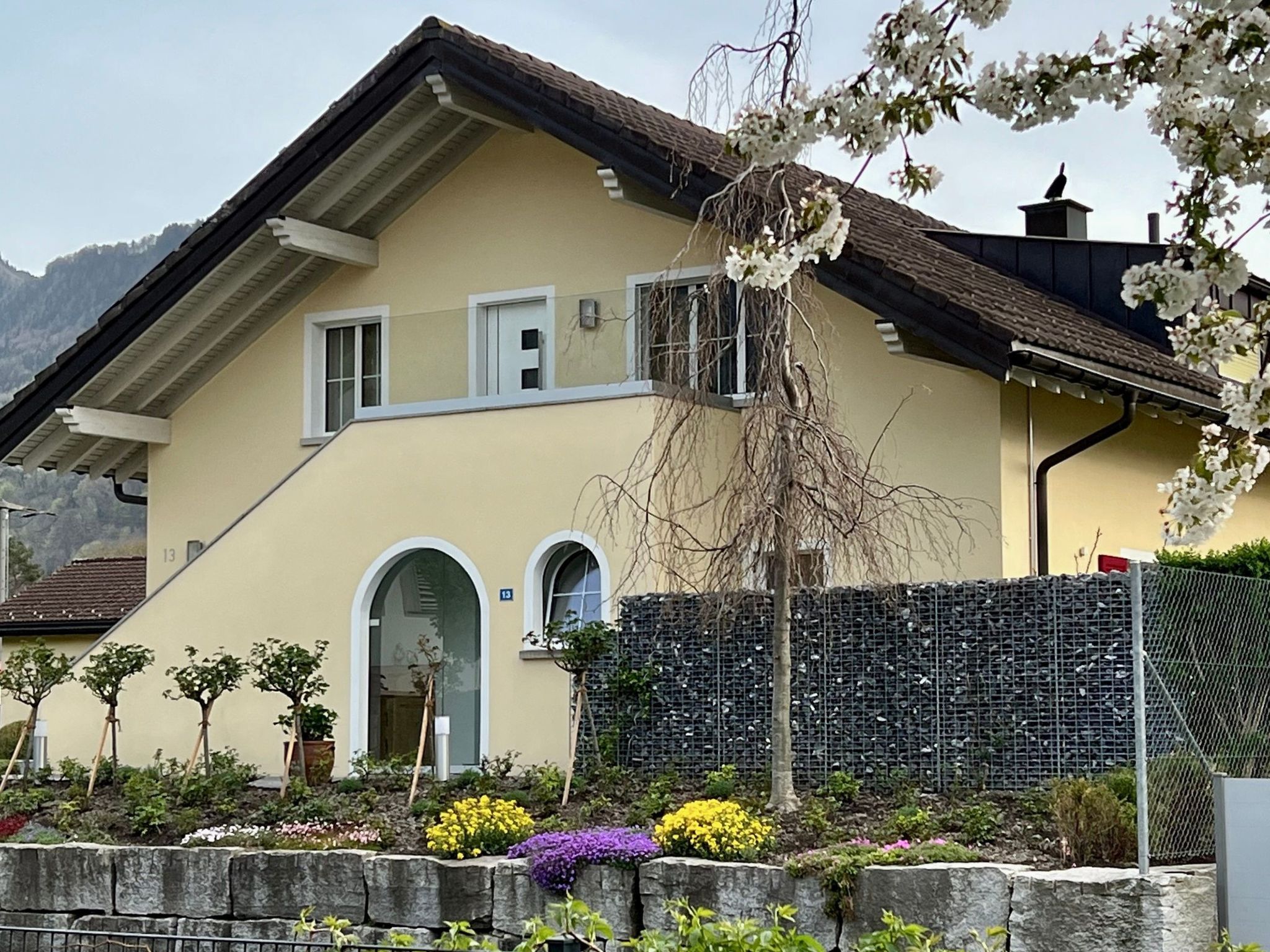 Uma casa de dois andares com telhado inclinado, parede de pedra e diversos plantas floridas no jardim frontal.
