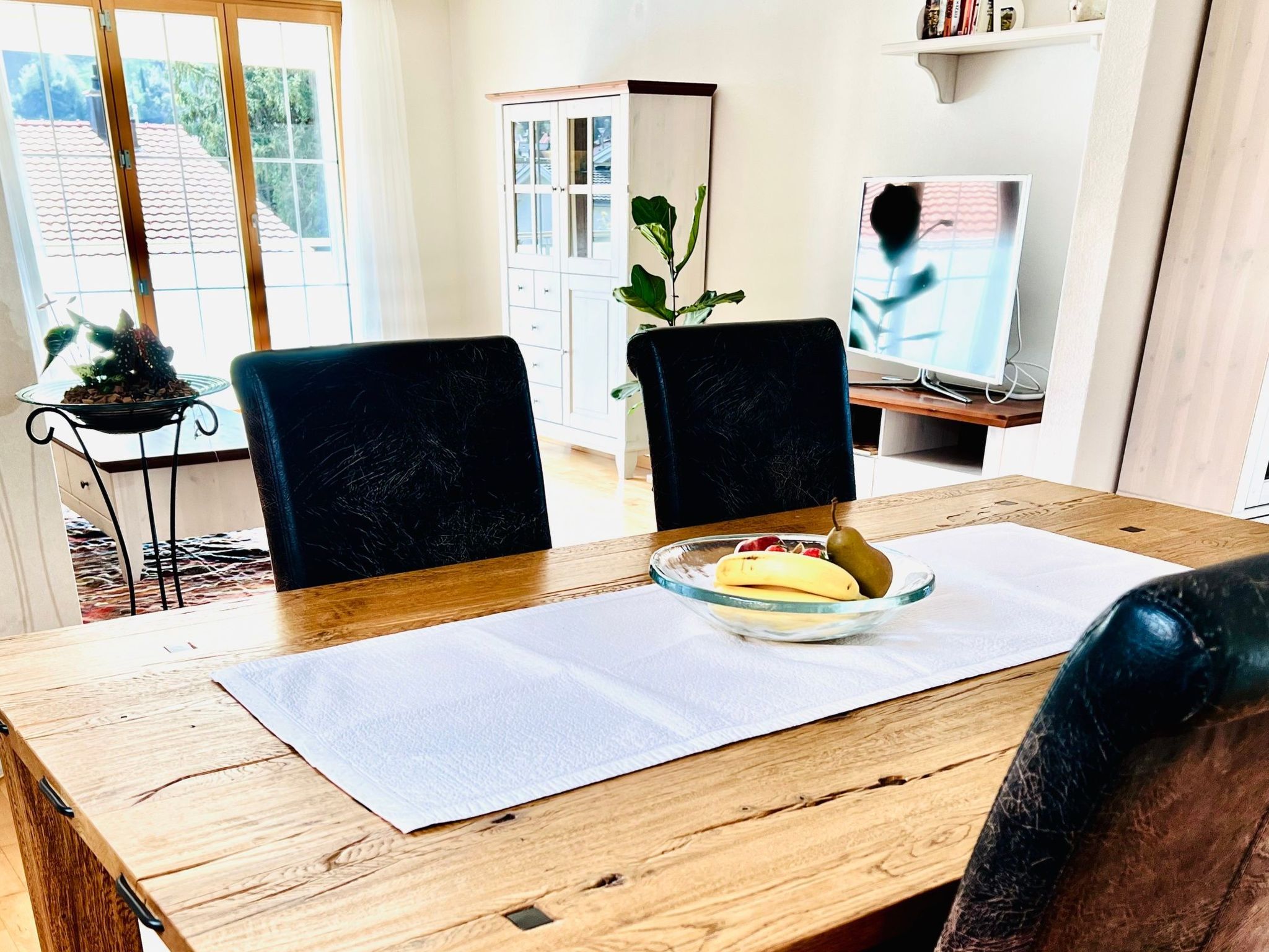 A dining area with a wooden table, two chairs, a bowl of fruit, and a television in the background.