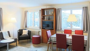 A modern living room with a sofa, a red ottoman, a television, a dining table with chairs, and a bookshelf. - Club Siesta Ferienwohnung/Studio, (Flumserberg Tannenheim), by Interhome (Flumserberg Tannenheim)