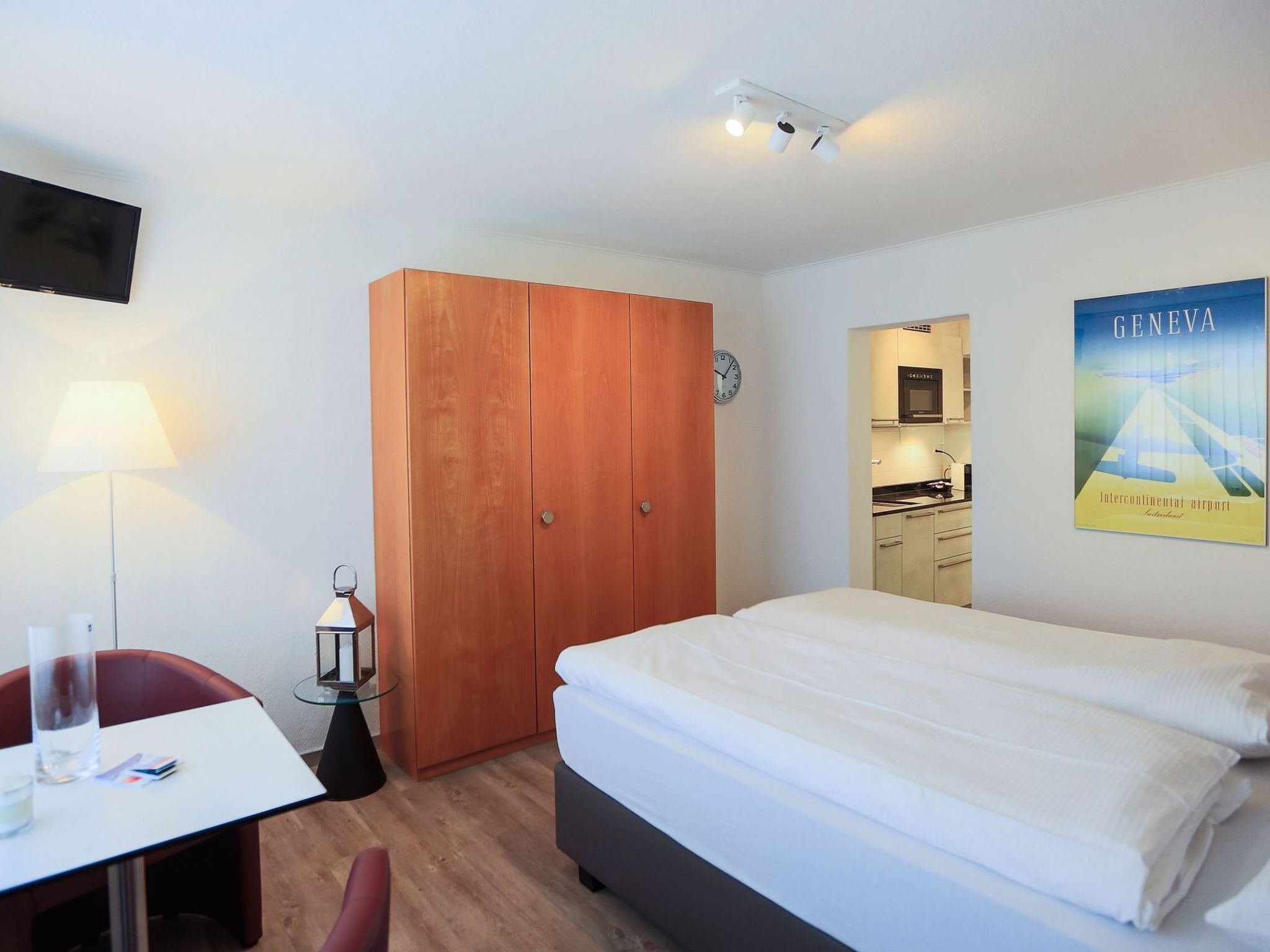 A hotel room with a bed, a desk with a chair, a wardrobe, a clock, and a poster on the wall.