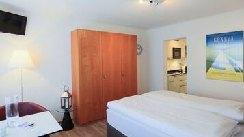 A hotel room with a bed, a desk with a chair, a wardrobe, a clock, and a poster on the wall.