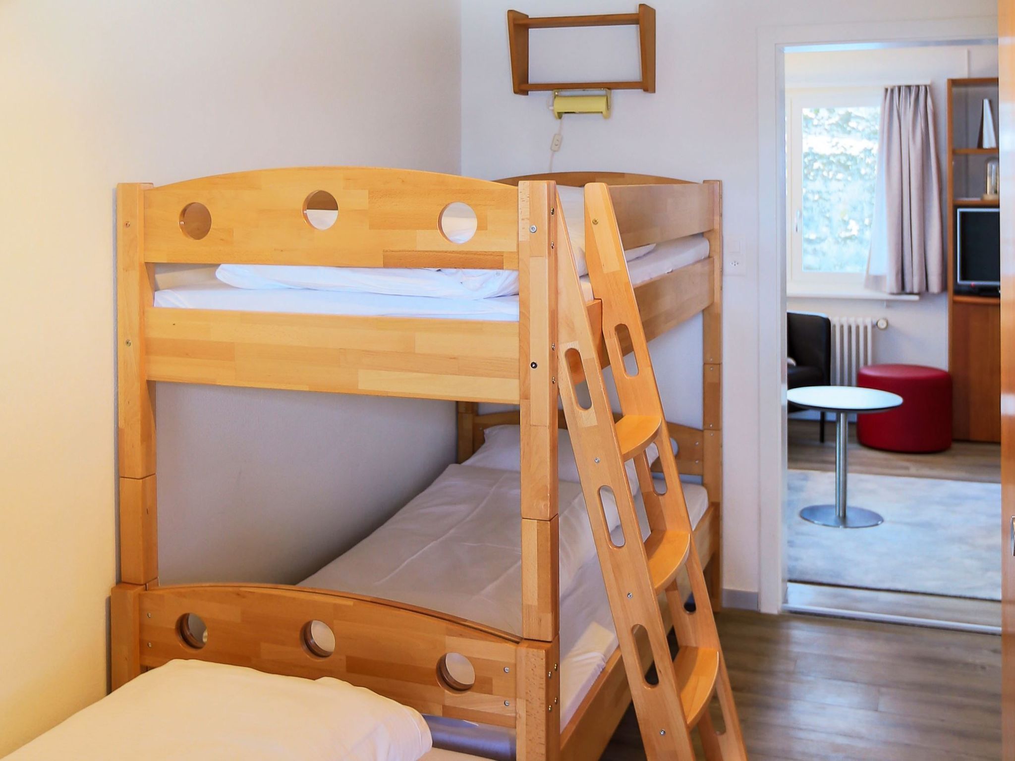 Una cama litera con escalera, una mesita pequeña y un televisor en una habitación con ventana y cortinas.
