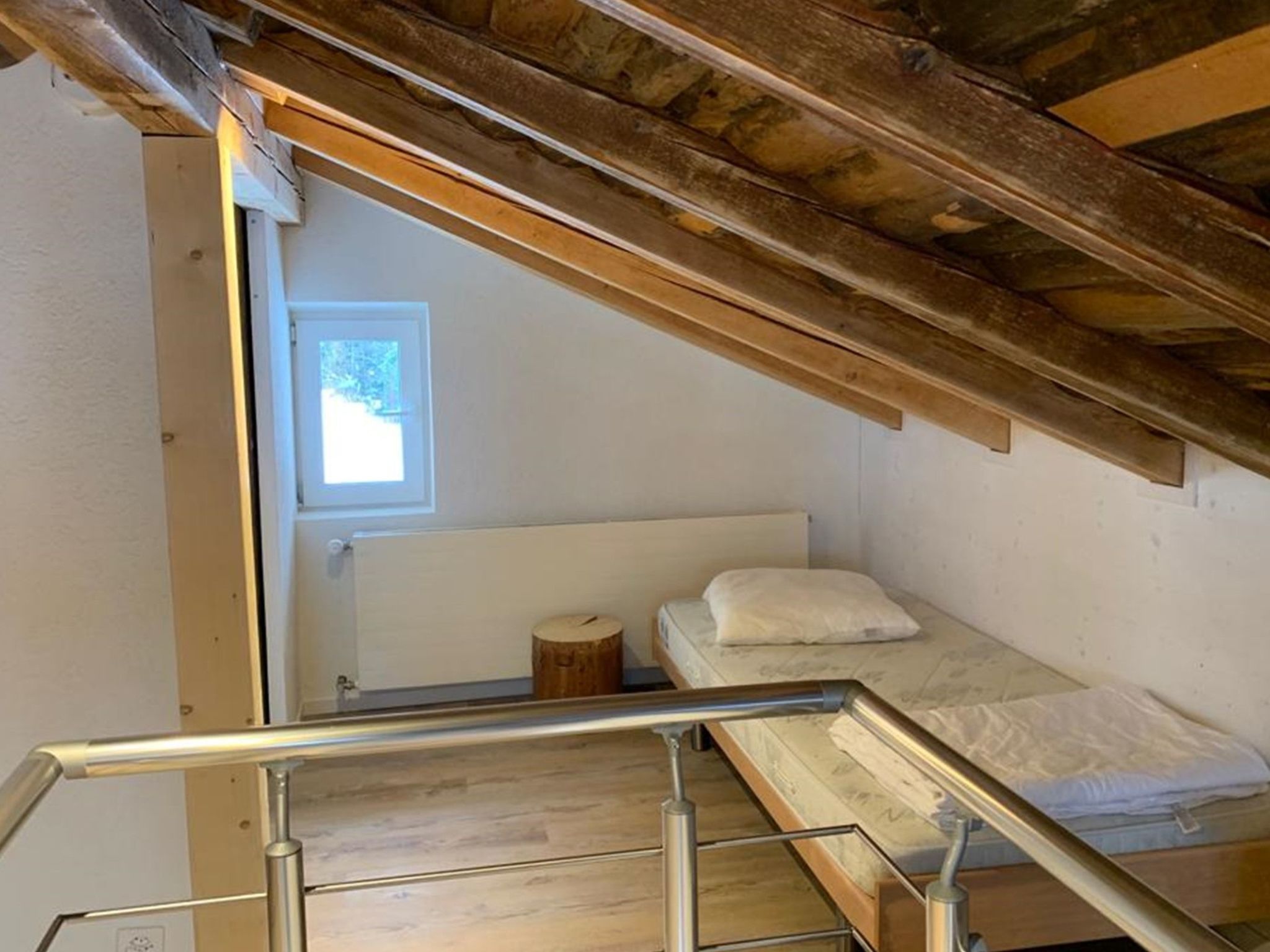 A compact room with a single bed, a small wooden stool, and a window providing natural light.