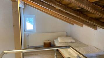 A compact room with a single bed, a small wooden stool, and a window providing natural light.