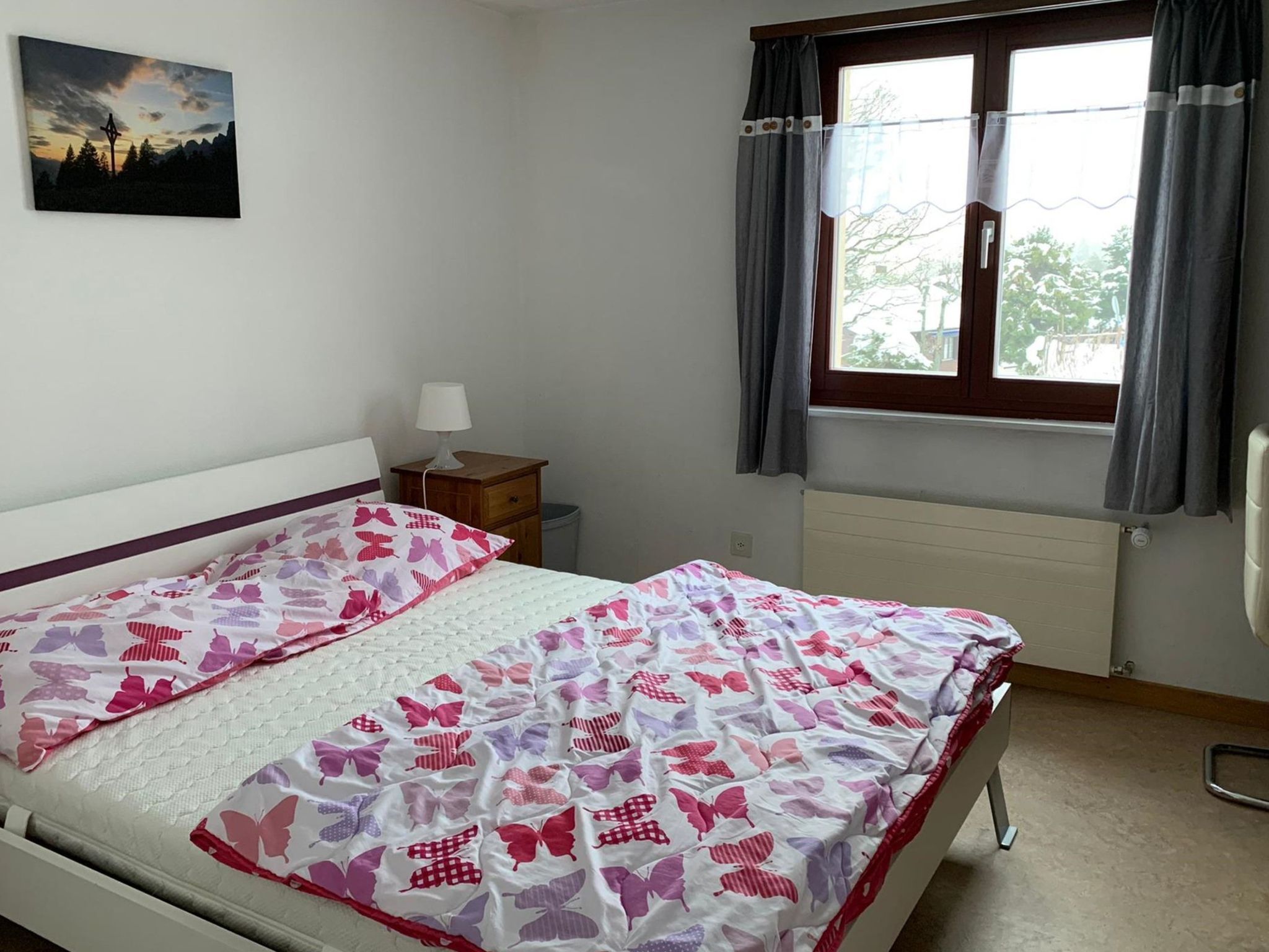 A bedroom with a bed, a nightstand, a lamp, a window with curtains, and a wall-mounted picture.