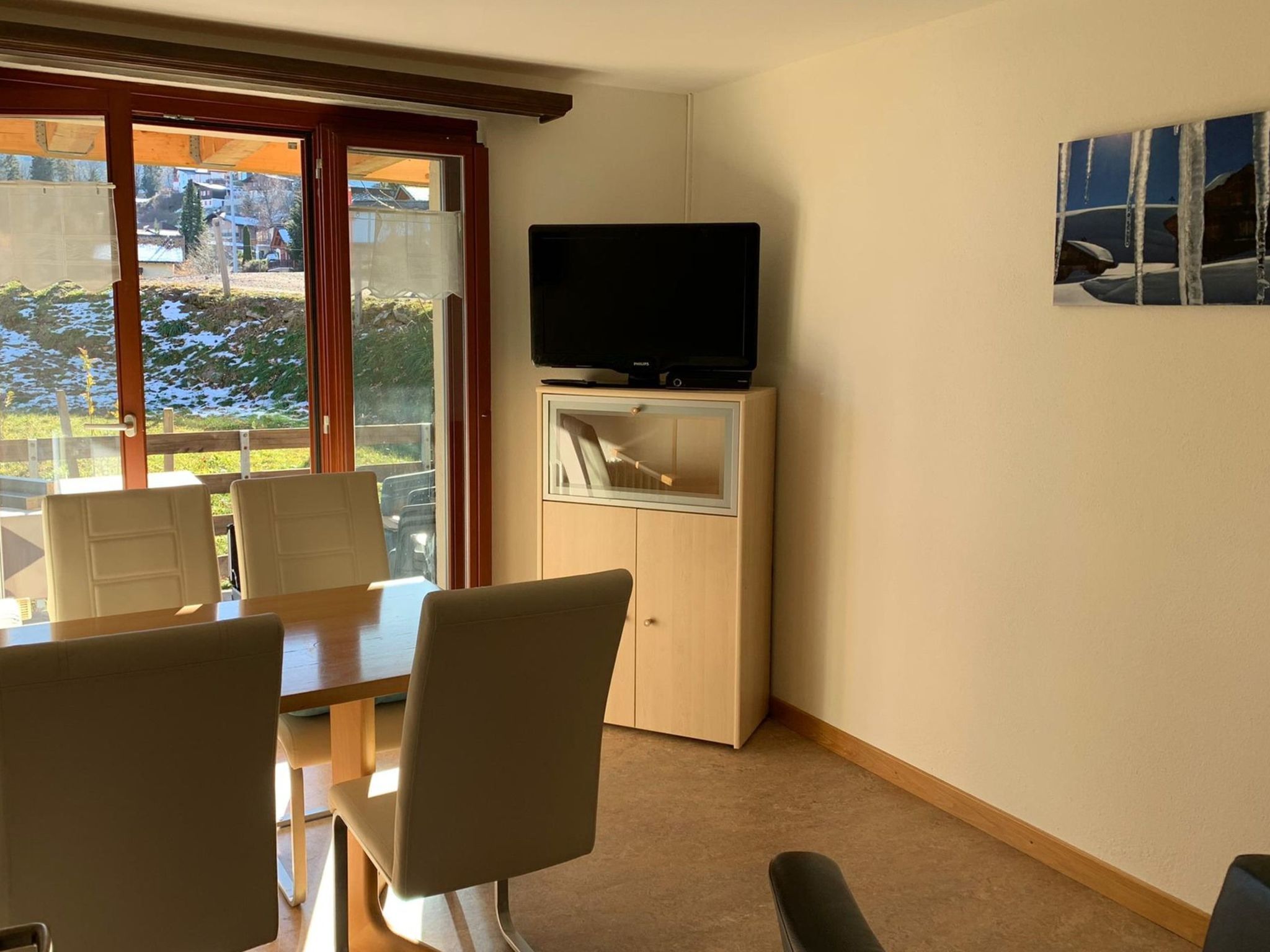 Ein Zimmer mit Esstisch, Stühlen, einem Fernseher auf einem Schrank und Blick durch eine Glastür.