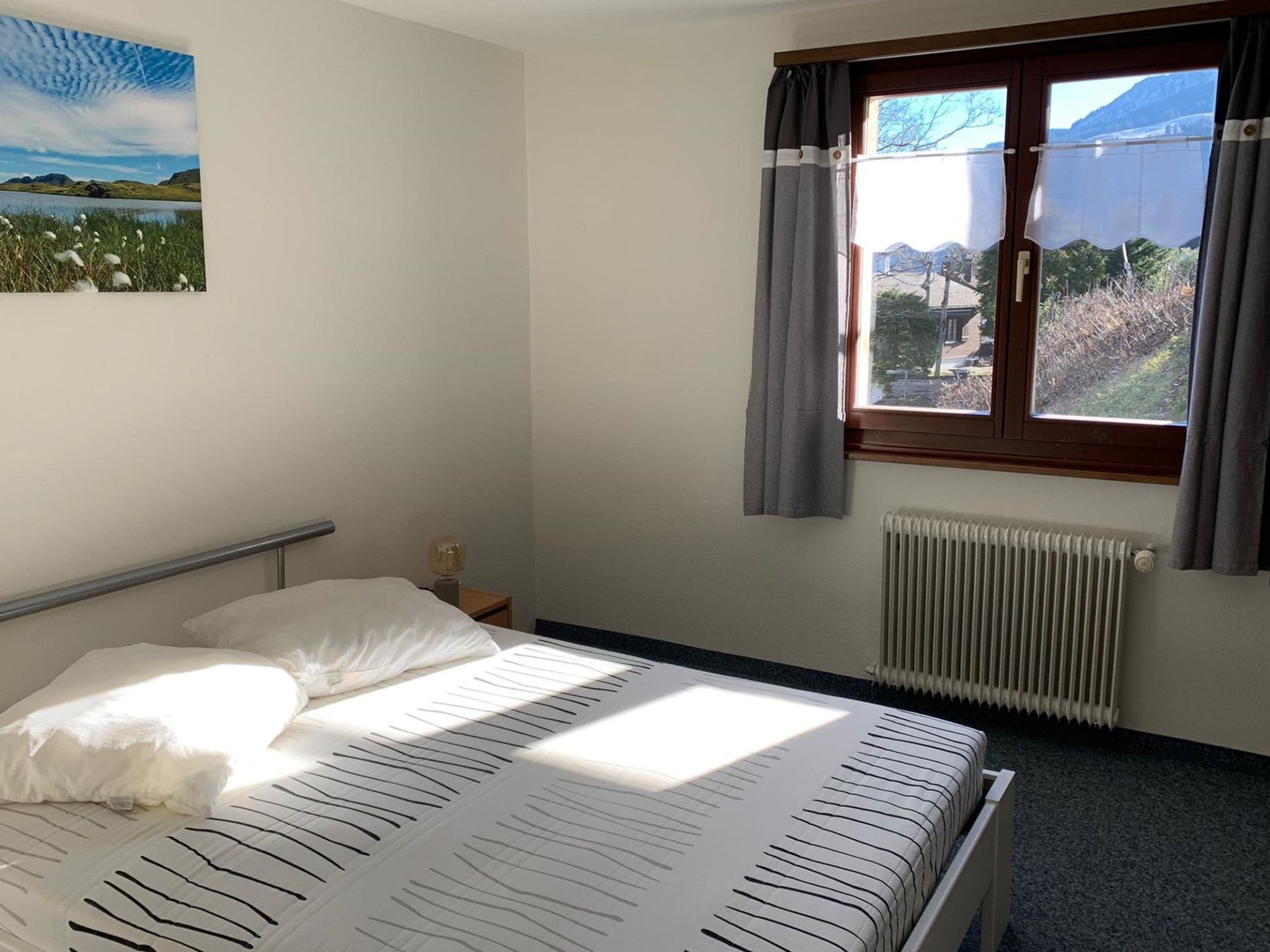 A bedroom with a bed, a window with curtains, a radiator, and a painting on the wall.