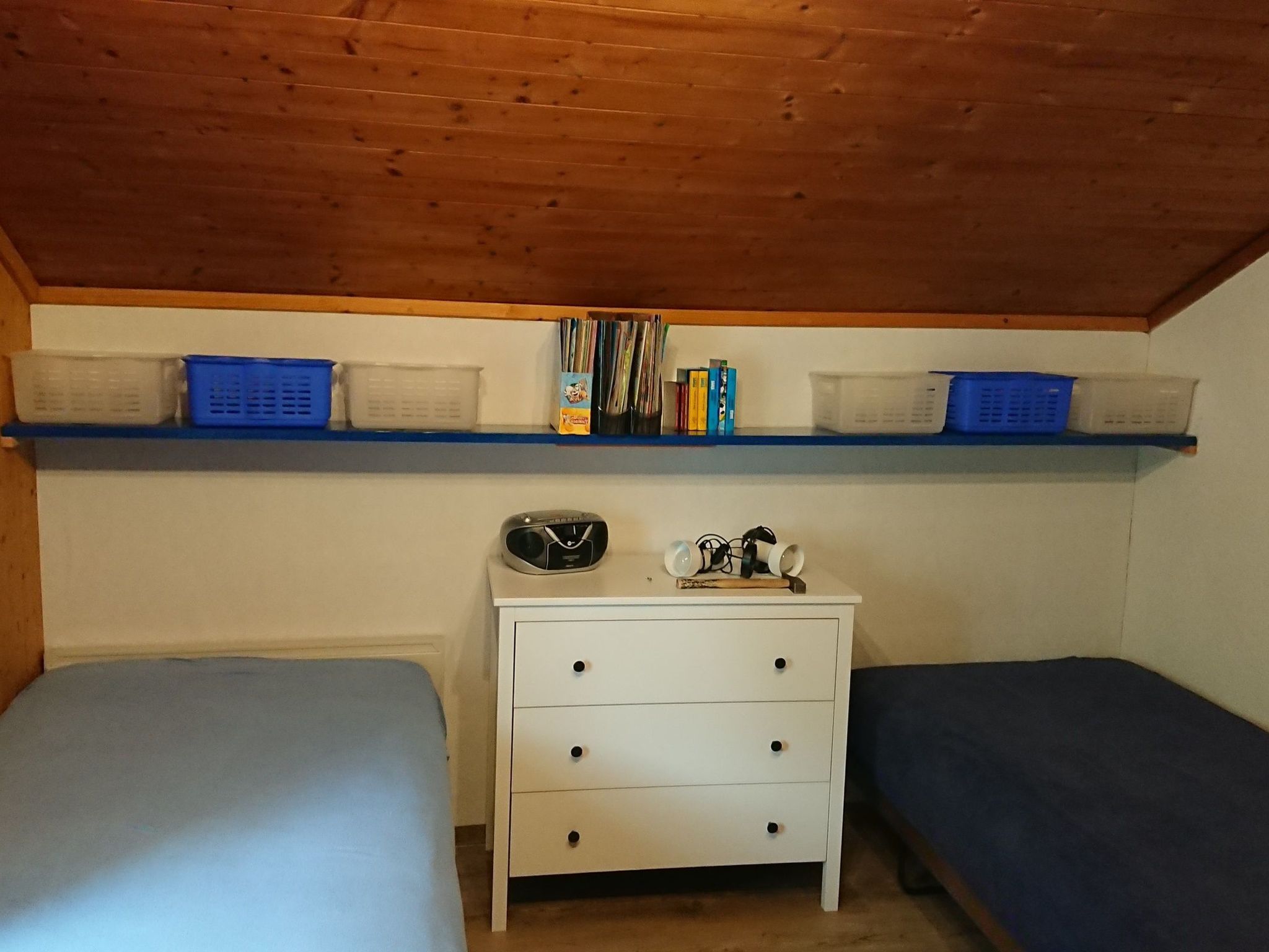 A small, enclosed room with two single beds, a white dresser with drawers, and shelves above holding various items including books and baskets.