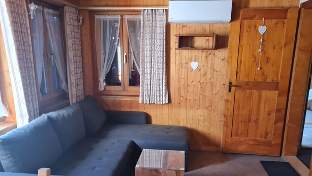 L’intérieur d’un chalet en bois, avec un canapé, une petite table en bois, une fenêtre avec des rideaux et une porte ornée d’un motif en forme de cœur.