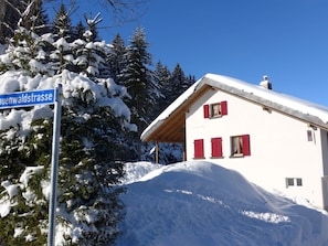 Ein schneebedecktes Gebäude mit einem roten Dach, einem strahlend blauen Himmel und einem Schild mit der Aufschrift 'Auenvaldstrasse'.