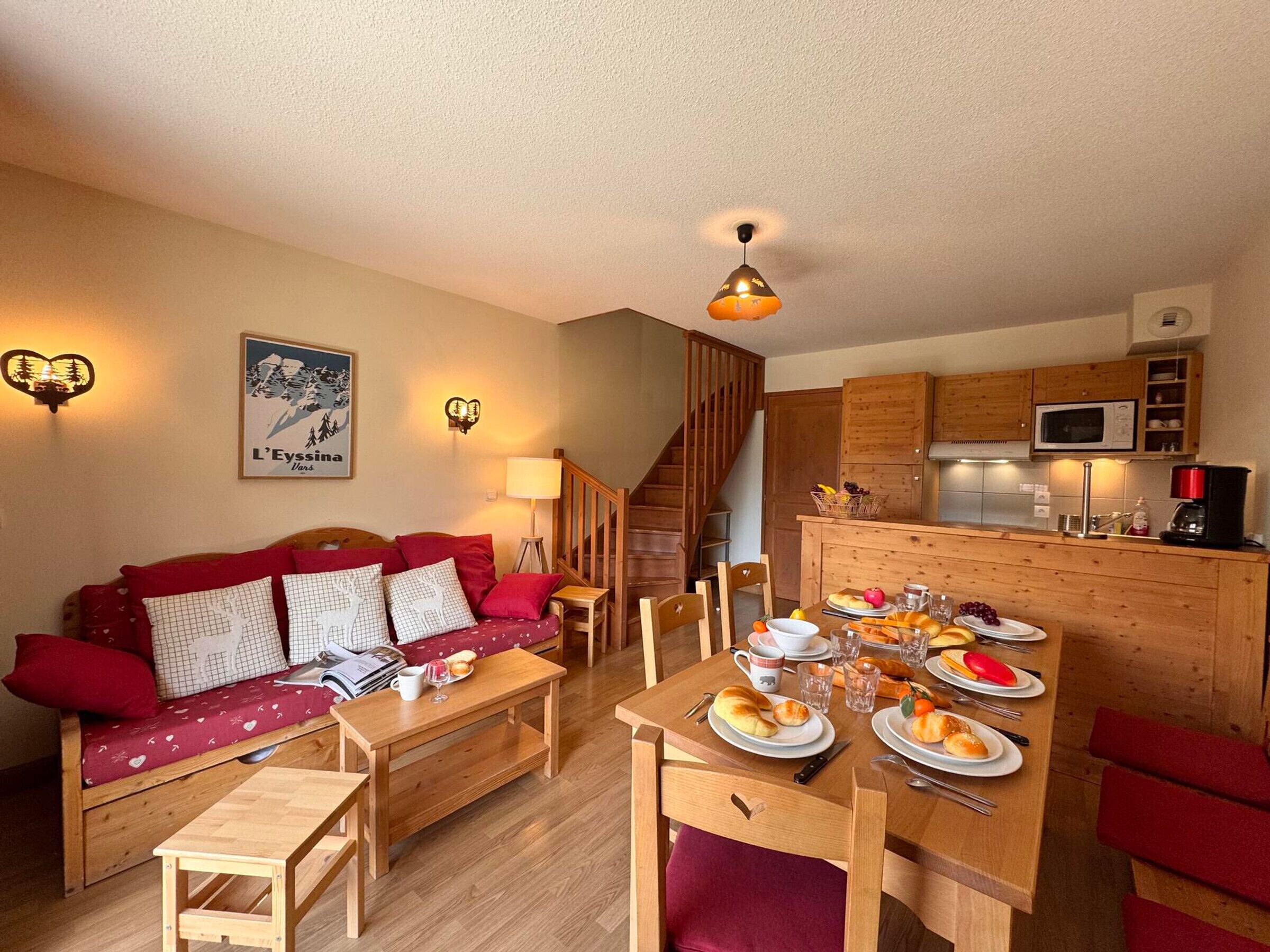 A cozy living area with a sofa, a dining table set with food, a kitchen with wooden cabinets, and a staircase.