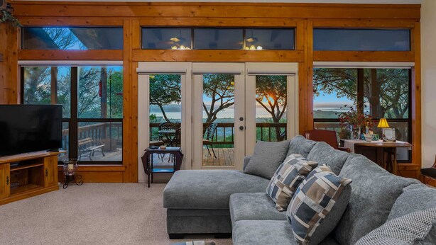 Una sala de estar acogedora con un sofá gris, un televisor de pantalla plana y ventanas amplias con vistas al exterior.