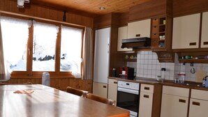 A wooden cabin interior with a dining table, kitchen area, and a window with a snowy view. - Chalet am Tobelbach, (Flumserberg Tannenheim), by Interhome (Flumserberg Tannenheim)