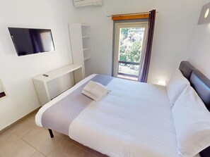 Ein ordentlich bezogenes Bett mit Kissen, ein flachbildfernseher an der Wand, ein kleiner Schreibtisch und ein Fenster mit Vorhängen.