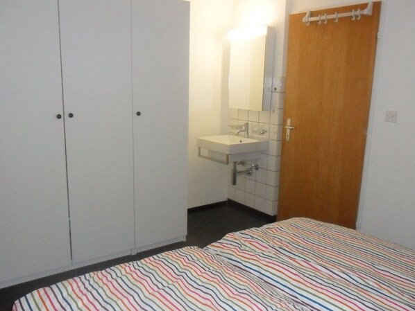 A bedroom with a bed, a wardrobe, a bathroom with a sink and mirror, and a door.