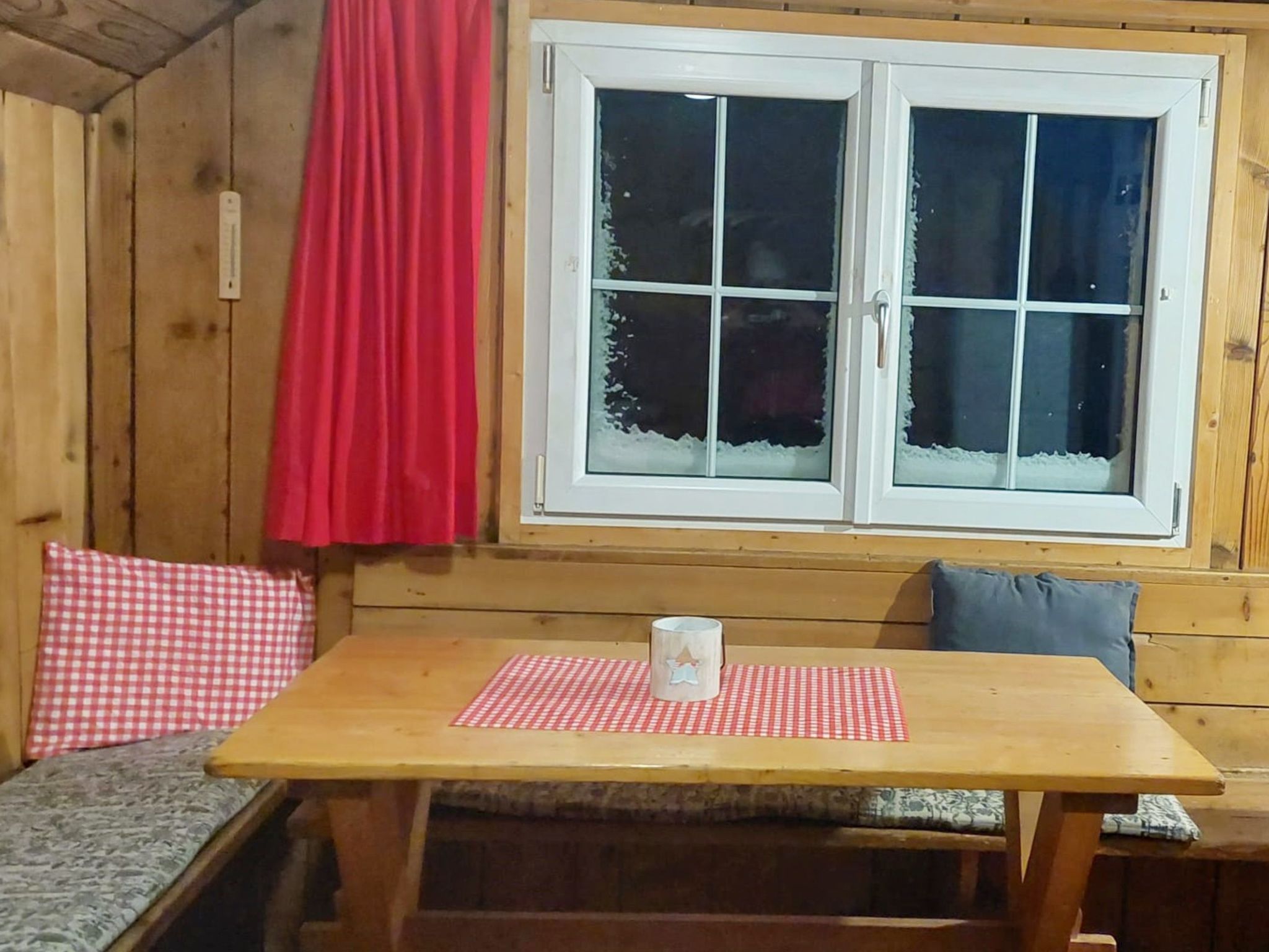A wooden cabin interior with a wooden table and bench, a window with frosted glass, and a red curtain.