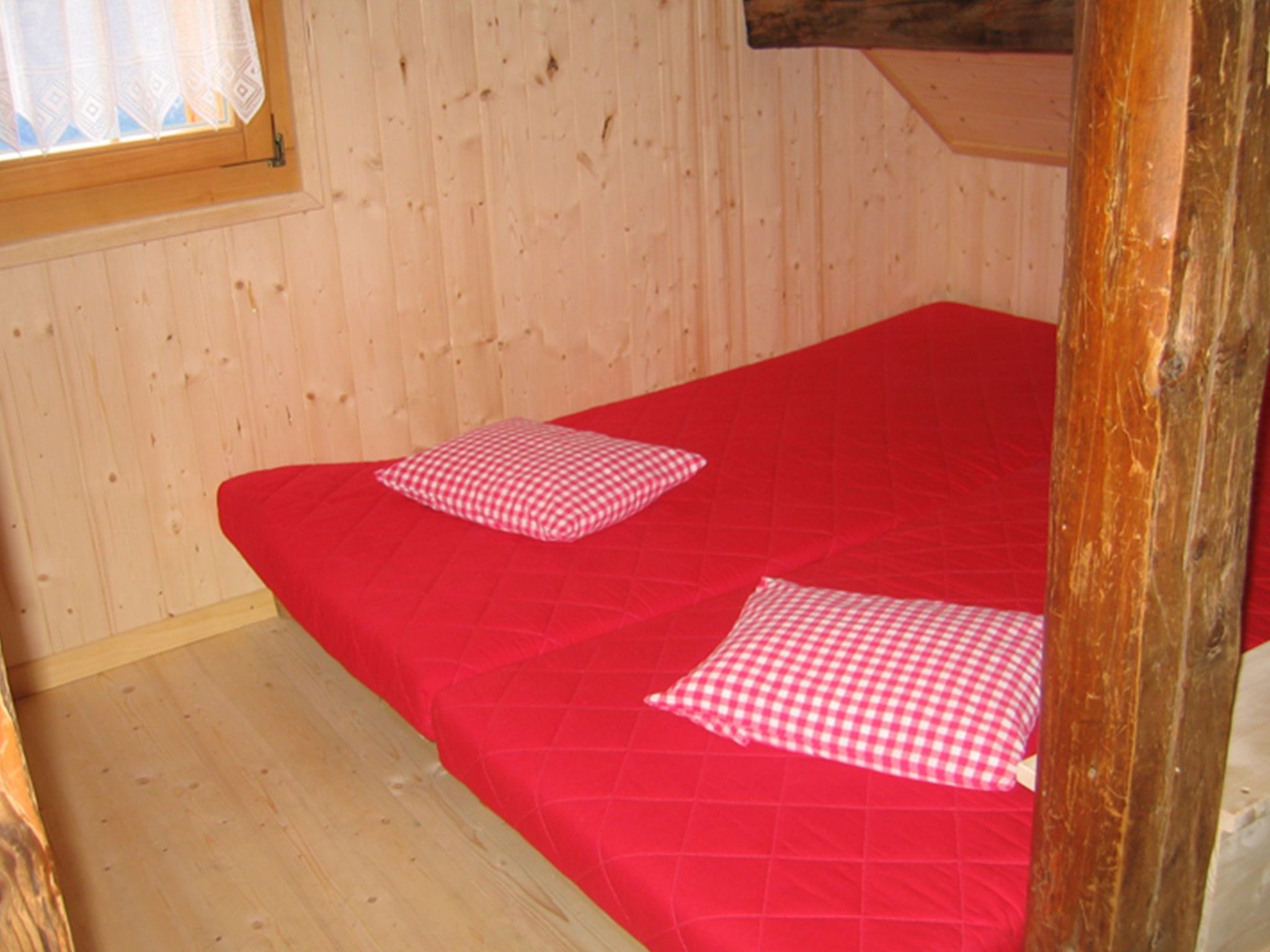 A single bed with a red bedspread and two checkered pillows in a room with wooden walls and a window with a white curtain.