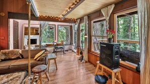 A cozy cabin interior with a wooden ceiling, large windows, a sofa, a dining table, and a speaker. - Woodland Home Near The Gorge - Private Deck 1 Bedroom Home (Washougal)