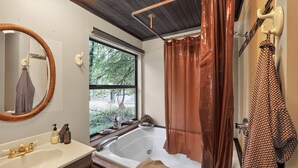 A bathroom with a bathtub, a window with a view of trees, a wooden ceiling, and a mirror with a towel hanging on it. - Woodland Home Near The Gorge - Private Deck 1 Bedroom Home (Washougal)