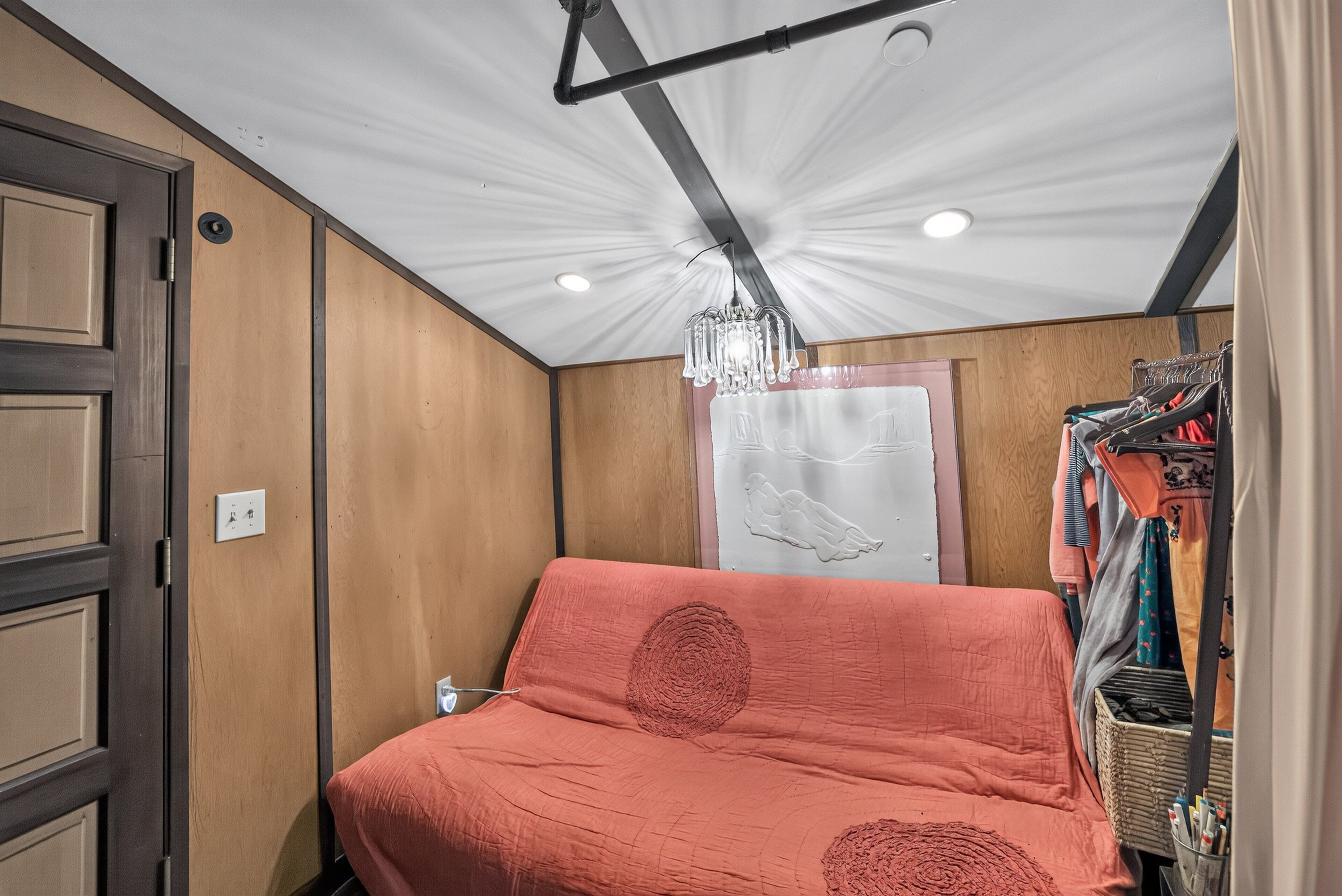 A compact room with a bed, a chandelier, a wardrobe, and a wall with a towel rack.