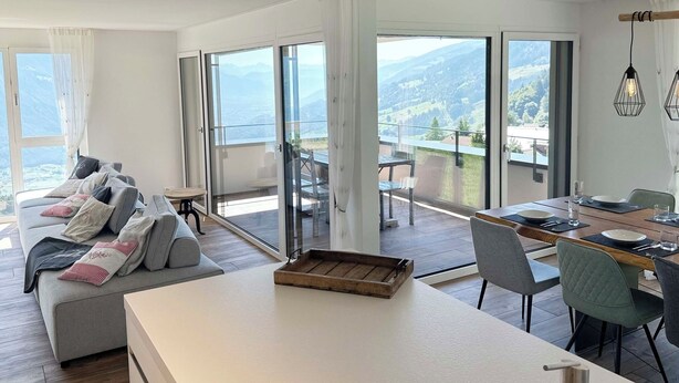 A modern living room with a sofa, a dining table, and a view of mountains through large glass doors.