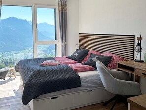 Ein Schlafzimmer mit Bett, Schreibtisch und Stuhl. Ein Fenster bietet Blick auf die Berge.