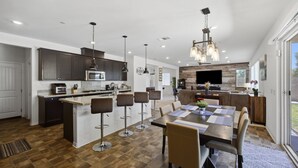 A modern kitchen with a central island, bar stools, a dining table with chairs, and a large TV. - Heritage Lake Luxury Home 4 Bedroom Home (Menifee)