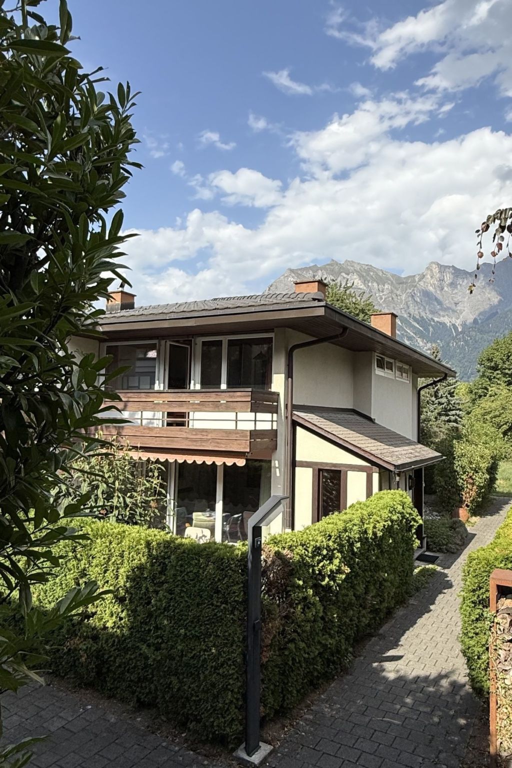 A two-story house with a balcony, surrounded by greenery and mountains in the background.
