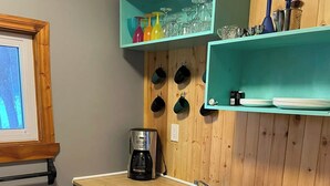 Una cocina con mostrador de madera, cafetera, lavabo y estantes con tazas y vasos.