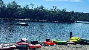Una playa con kayaks y una pequeña embarcación en la arena, cielo despejado y un avión volando en el horizonte.