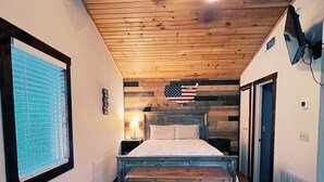 Una camera da letto con soffitto in legno, un letto con testiera in legno, una finestra con persiane e una bandiera americana appesa al muro.