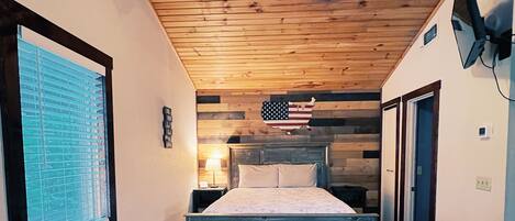 A bedroom with a wooden ceiling, a bed with a wooden headboard, a window with blinds, and a wall-mounted American flag.