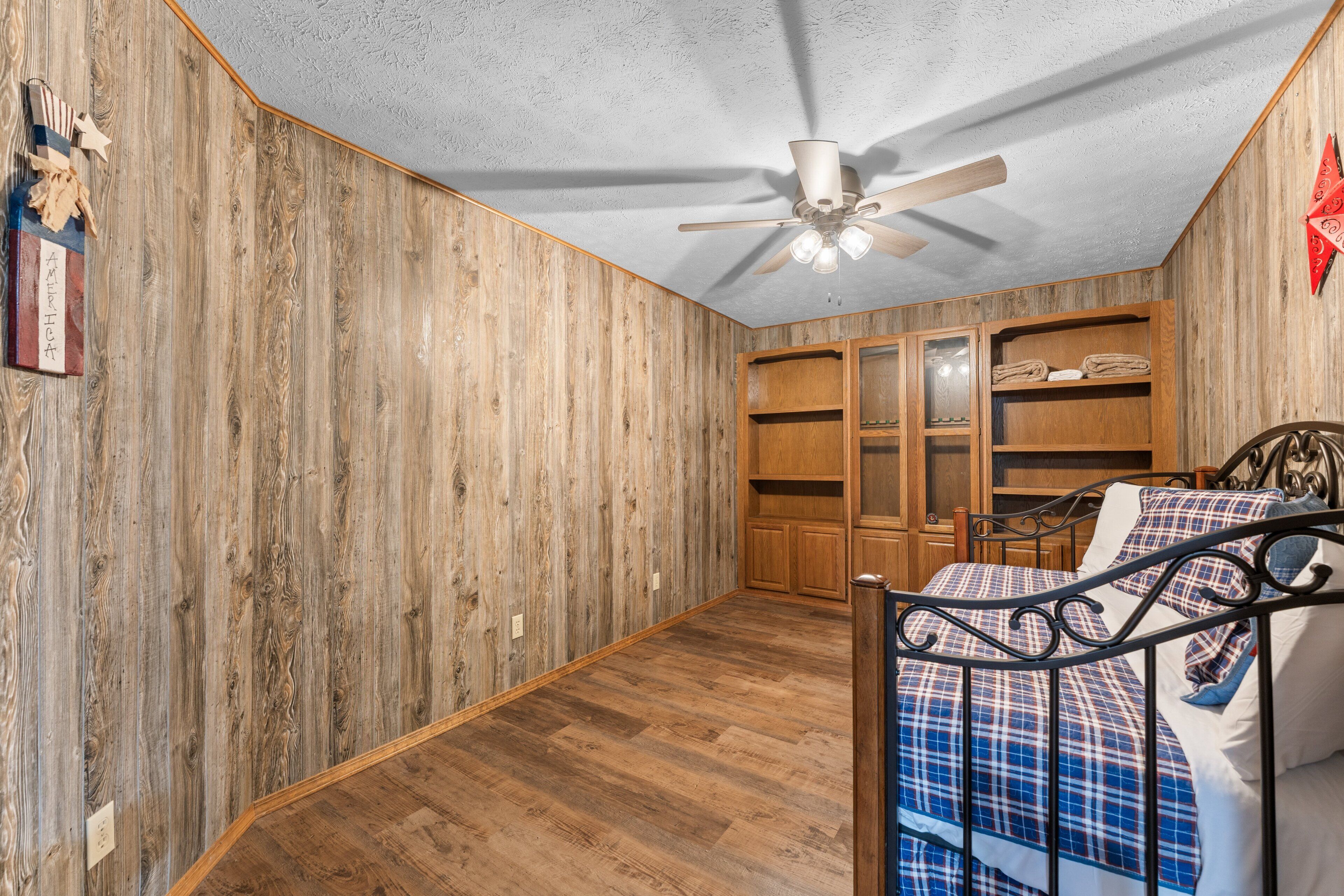 A bedroom with wooden walls, a ceiling fan, a bed with a plaid bedspread, and built-in wooden shelving.