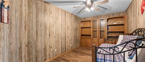 A bedroom with wooden walls, a ceiling fan, a bed with a plaid bedspread, and built-in wooden shelving.