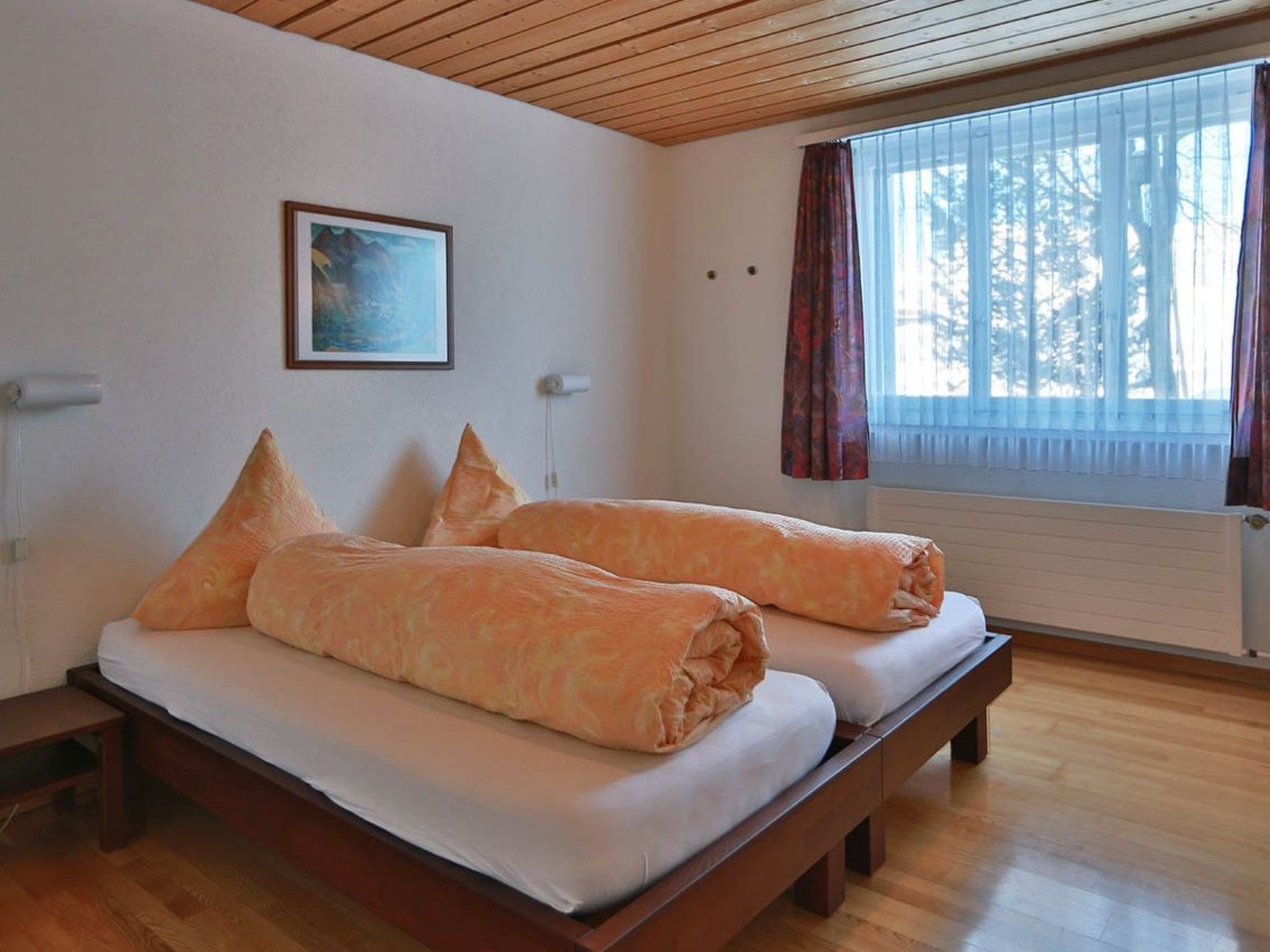 Ein Zimmer mit Holzboden, einem Bett mit Kissen, einem Bild an der Wand und einem Fenster mit Vorhängen.