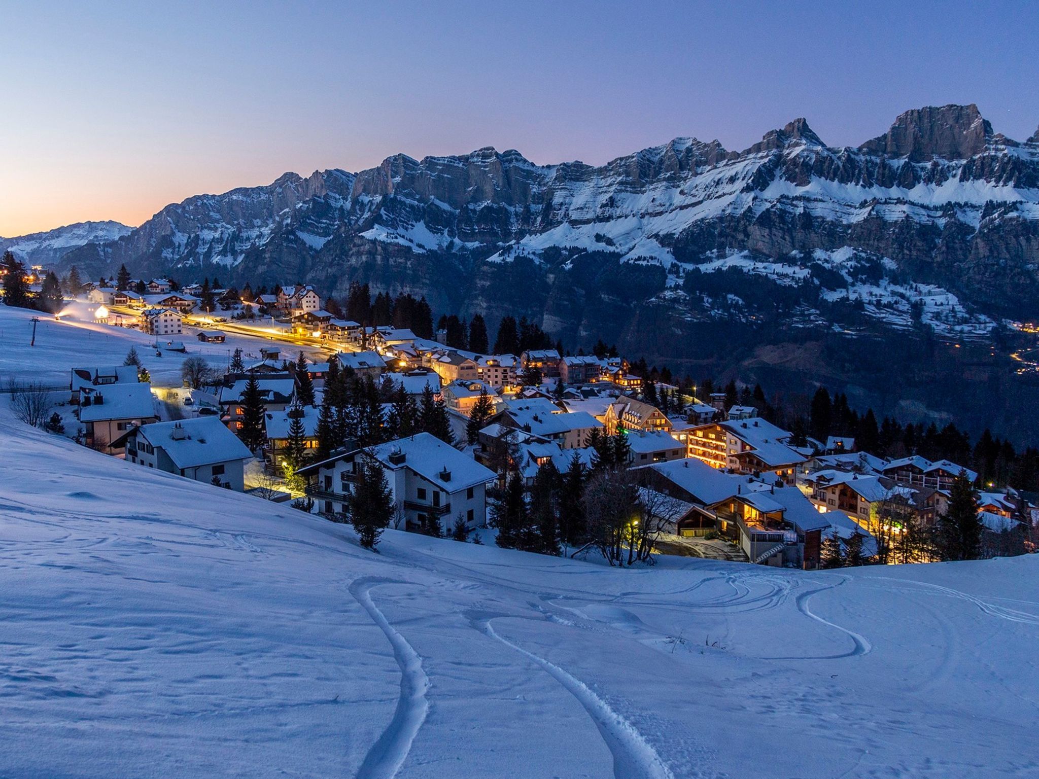 A snow-covered mountain village at dusk with illuminated buildings and ski tracks.