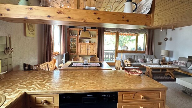 A wooden interior with a kitchen, dining area, and living space, featuring a dishwasher, wooden cabinets, and a sofa.