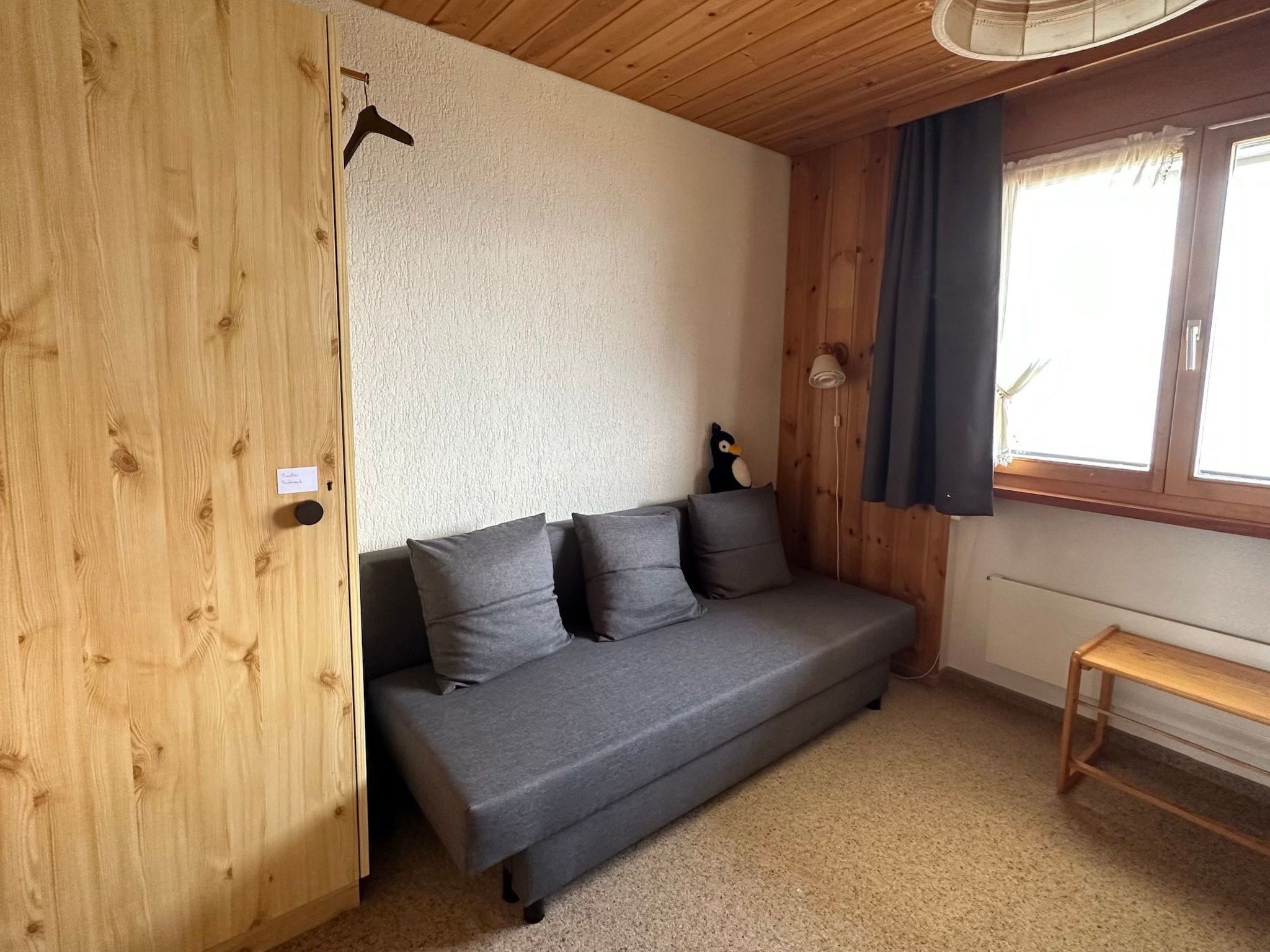 A room with a wooden wardrobe, a grey sofa with cushions, a small table, and a window with curtains.