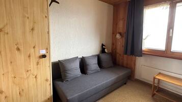 A room with a wooden wardrobe, a grey sofa with cushions, a small table, and a window with curtains.