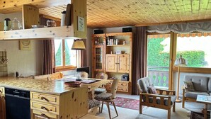 A cozy cabin interior with a wooden dining table, chairs, a kitchen area with a dishwasher, and a large window offering a view of greenery.